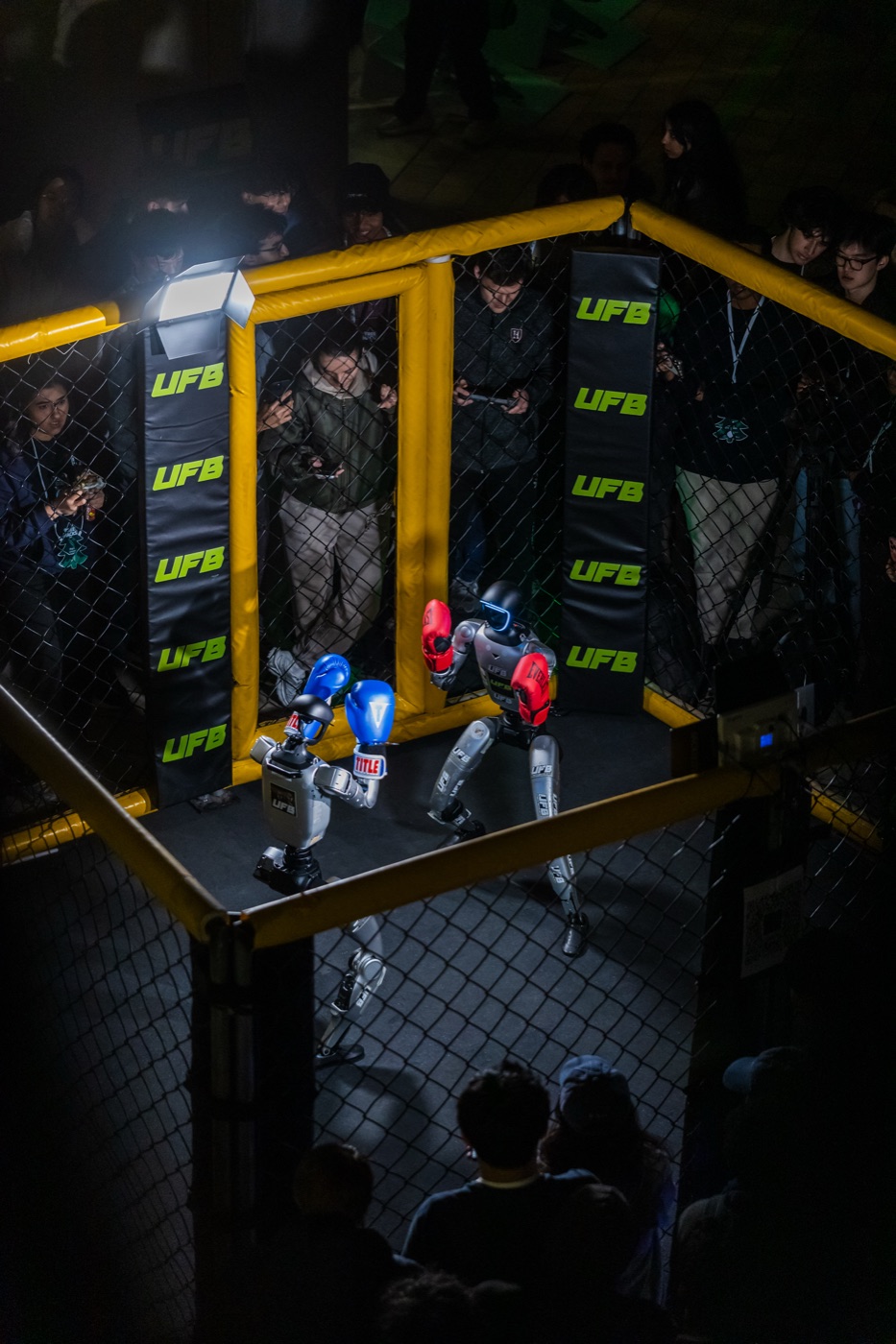 Two humanoid robots boxing inside the UFB cage at TreeHacks 2026, run by hackers with handheld controllers at the fence
